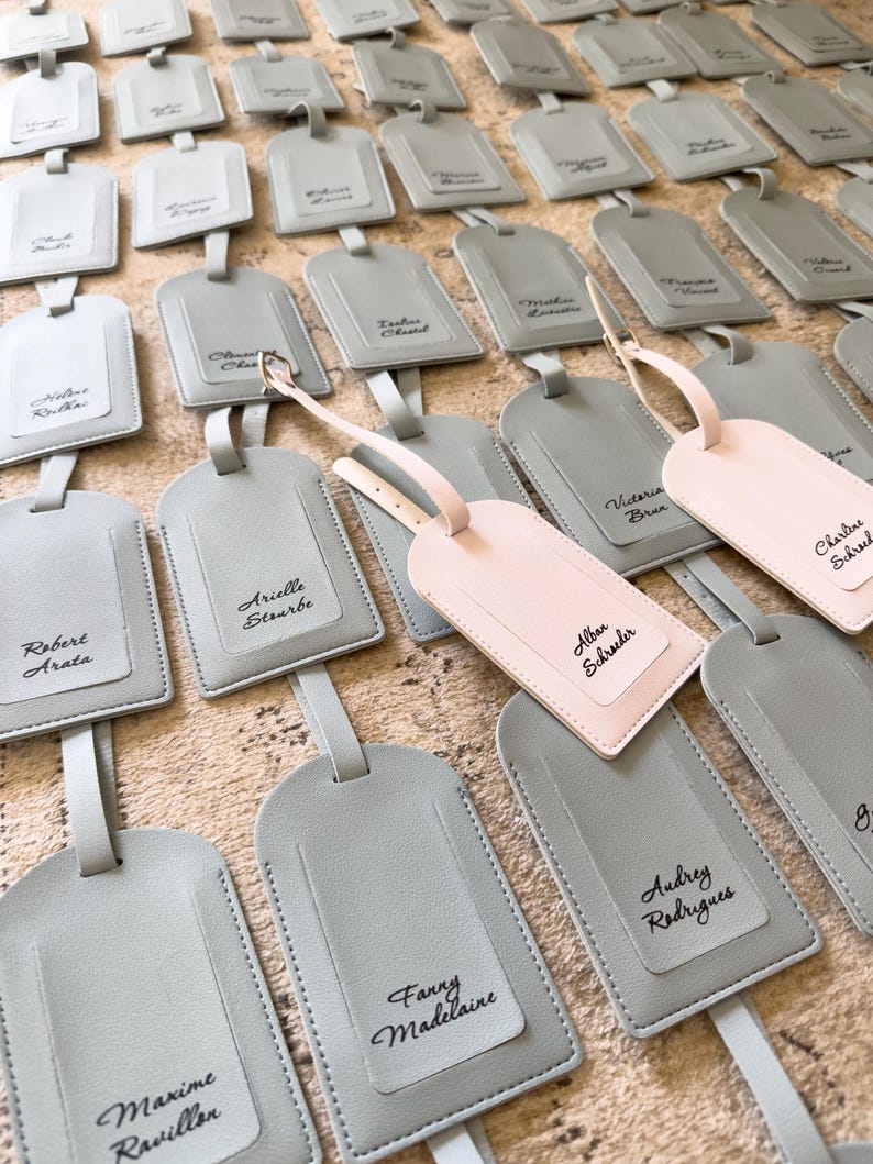 May include: A selection of personalised luggage tags in shades of grey and one in pink. Each tag has a name written in script. The tags are made of a smooth material and have a strap for attachment.