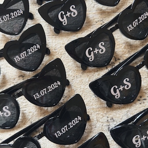 May include: Black heart-shaped sunglasses with the initials "G+S" and the date "13.07.2024" printed in white. The sunglasses have dark lenses and a classic frame design, perfect for a wedding or special event.
