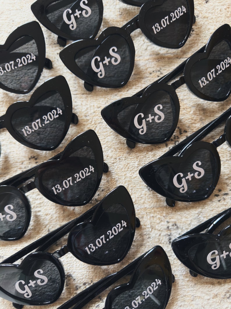 May include: Black heart-shaped sunglasses with the initials "G+S" and the date "13.07.2024" printed in white. The sunglasses have dark lenses and a classic frame design, perfect for a wedding or special event.