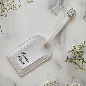 May include: A white luggage tag with a silver buckle and strap. The tag features the name "Laura Rossi" in black script. The tag is set against a marble background with delicate white flowers.