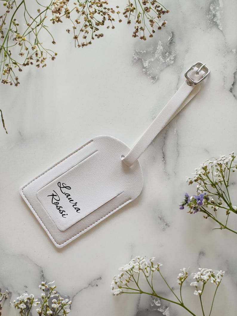 May include: A white luggage tag with a silver buckle and strap. The tag features the name "Laura Rossi" in black script. The tag is set against a marble background with delicate white flowers.