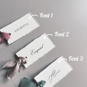 May include: Three white rectangular place cards with torn edges, each featuring a different name in elegant black script. The cards are adorned with silk ribbons in shades of pink and green, set against a gray background.