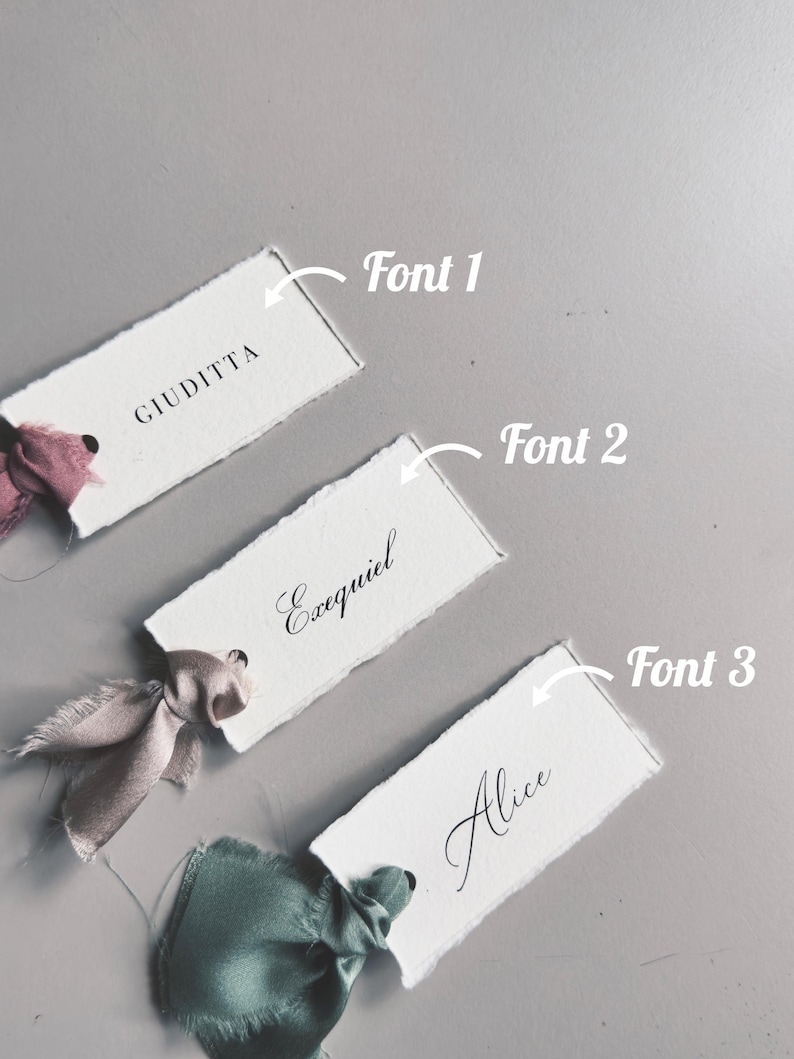 May include: Three white rectangular place cards with torn edges, each featuring a different name in elegant black script. The cards are adorned with silk ribbons in shades of pink and green, set against a gray background.