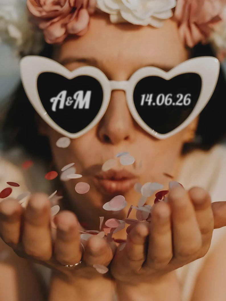 May include: White heart-shaped sunglasses with black lenses, featuring the initials "A&M" and the date "14.06.26". The person is blowing confetti, with a floral crown in the background. The confetti is in shades of pink and red.