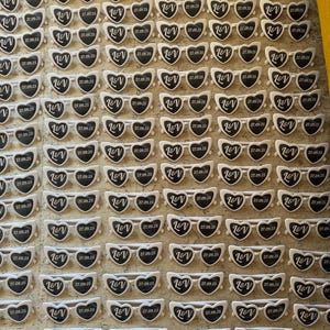 May include: A grid of heart-shaped sunglasses with white frames and black lenses. Each pair has the initials "L&V" and the date "27.09.25" printed on them. The sunglasses are arranged on a textured surface.