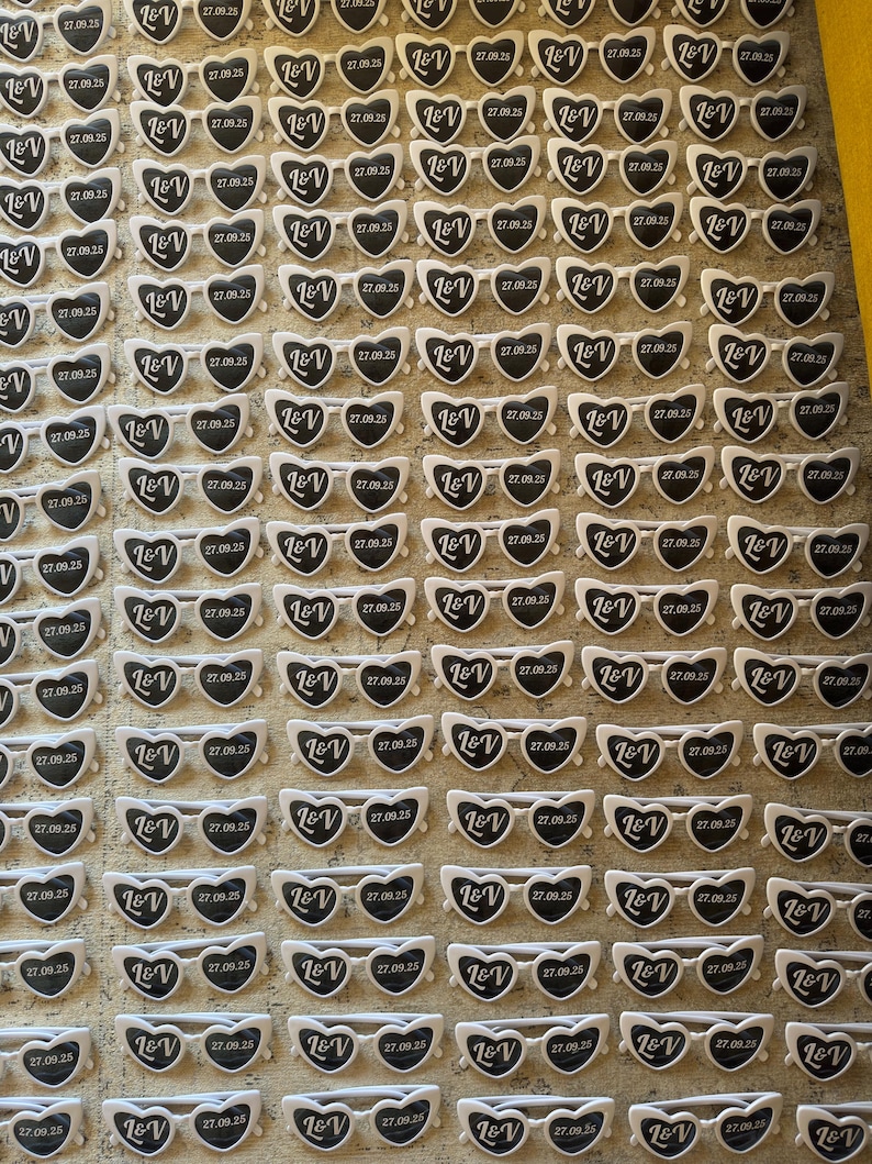 May include: A grid of heart-shaped sunglasses with white frames and black lenses. Each pair has the initials "L&V" and the date "27.09.25" printed on them. The sunglasses are arranged on a textured surface.
