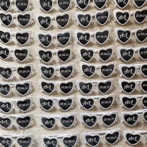 May include: Rows of white heart-shaped sunglasses with black lenses are arranged on a textured surface. Each pair features the initials "A&L" and the date "07.09.24" printed on the lenses, suggesting a wedding or event theme.