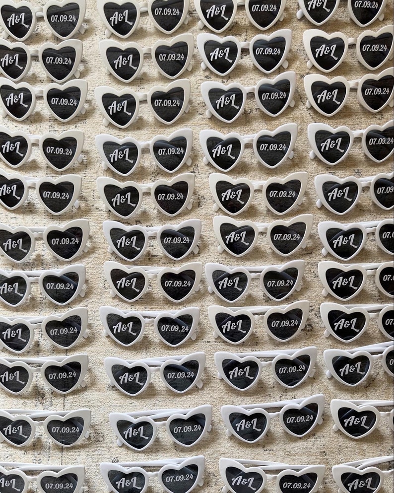May include: Rows of white heart-shaped sunglasses with black lenses are arranged on a textured surface. Each pair features the initials "A&L" and the date "07.09.24" printed on the lenses, suggesting a wedding or event theme.