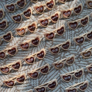 May include: Rows of heart-shaped sunglasses with cream-colored frames and tinted lenses. Each pair has the text "M&M" and "01.07.26" printed on the lenses. The sunglasses are arranged on a textured surface.