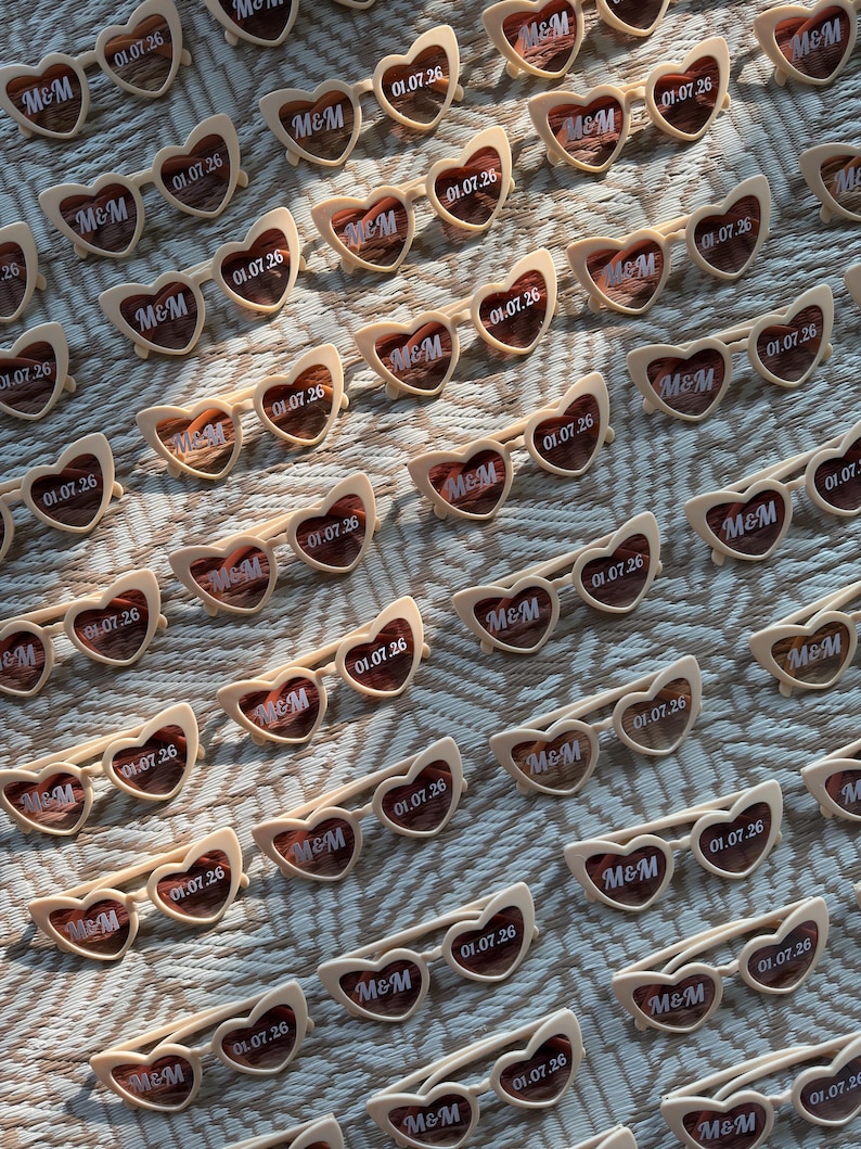 May include: Rows of heart-shaped sunglasses with cream-colored frames and tinted lenses. Each pair has the text "M&M" and "01.07.26" printed on the lenses. The sunglasses are arranged on a textured surface.