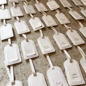 May include: An array of off-white luggage tags with attached straps, neatly arranged. Each tag has a smaller, rectangular insert with a name printed in gold. The tags are displayed on a textured, neutral-toned surface.