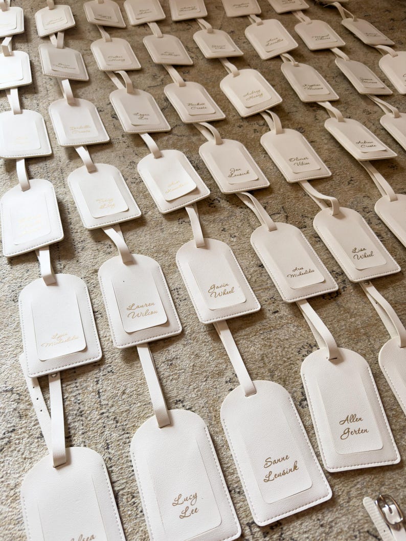 May include: An array of off-white luggage tags with attached straps, neatly arranged. Each tag has a smaller, rectangular insert with a name printed in gold. The tags are displayed on a textured, neutral-toned surface.