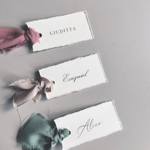 May include: Three handmade place cards with torn edges, each featuring a name in elegant calligraphy. The cards are adorned with silk ribbons in shades of pink and green, adding a touch of sophistication. Perfect for weddings or special events.
