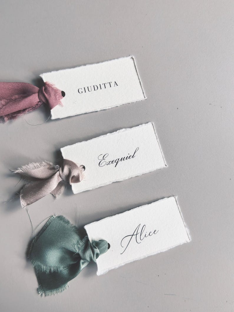May include: Three handmade place cards with torn edges, each featuring a name in elegant calligraphy. The cards are adorned with silk ribbons in shades of pink and green, adding a touch of sophistication. Perfect for weddings or special events.