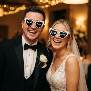May include: A smiling couple at a wedding, wearing heart-shaped sunglasses. The sunglasses are white with black text that reads "L&A" and "05.06.26". The man wears a black tuxedo with a bow tie and a white rose boutonniere. The woman wears a white lace wedding dress.