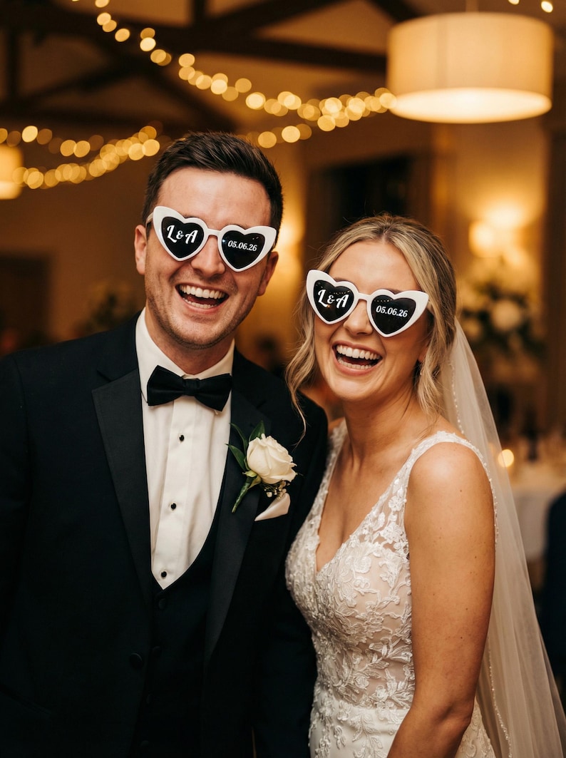 May include: A smiling couple at a wedding, wearing heart-shaped sunglasses. The sunglasses are white with black text that reads "L&A" and "05.06.26". The man wears a black tuxedo with a bow tie and a white rose boutonniere. The woman wears a white lace wedding dress.