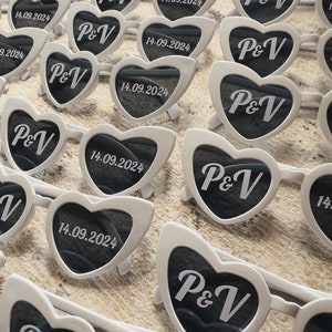 May include: White heart-shaped sunglasses with dark lenses, featuring the initials "P&V" and the date "14.09.2024" printed on the lenses. The sunglasses are arranged on a textured surface, suggesting a wedding or event theme.