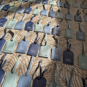 May include: A selection of leather luggage tags in shades of blue and grey, displayed on a textured surface. Each tag features a strap and buckle, with names printed in a delicate font. The tags vary in colour from light to dark.