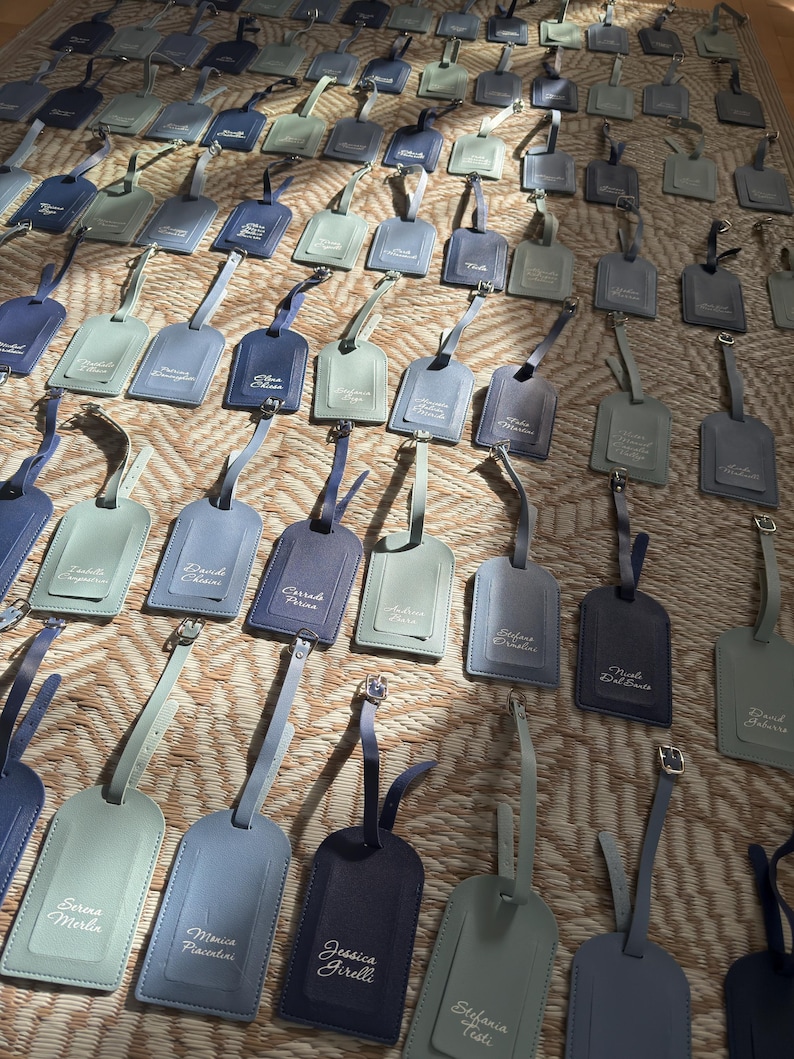 May include: A selection of leather luggage tags in shades of blue and grey, displayed on a textured surface. Each tag features a strap and buckle, with names printed in a delicate font. The tags vary in colour from light to dark.