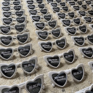 May include: Rows of white heart-shaped sunglasses with dark lenses are arranged on a textured surface. Each pair features the text "Martuzzo Wedding" and the date "26.06.24". The sunglasses have white frames and are likely wedding favors.