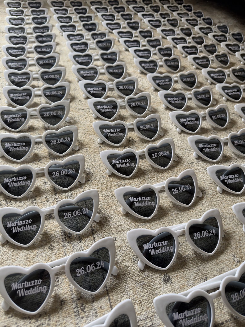 May include: Rows of white heart-shaped sunglasses with dark lenses are arranged on a textured surface. Each pair features the text "Martuzzo Wedding" and the date "26.06.24". The sunglasses have white frames and are likely wedding favors.