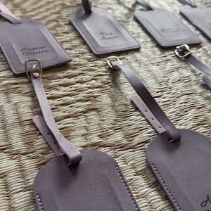 May include: Lavender luggage tags with personalized names in black script. Each tag has a matching strap with a gold-coloured buckle. The tags are arranged on a woven surface.