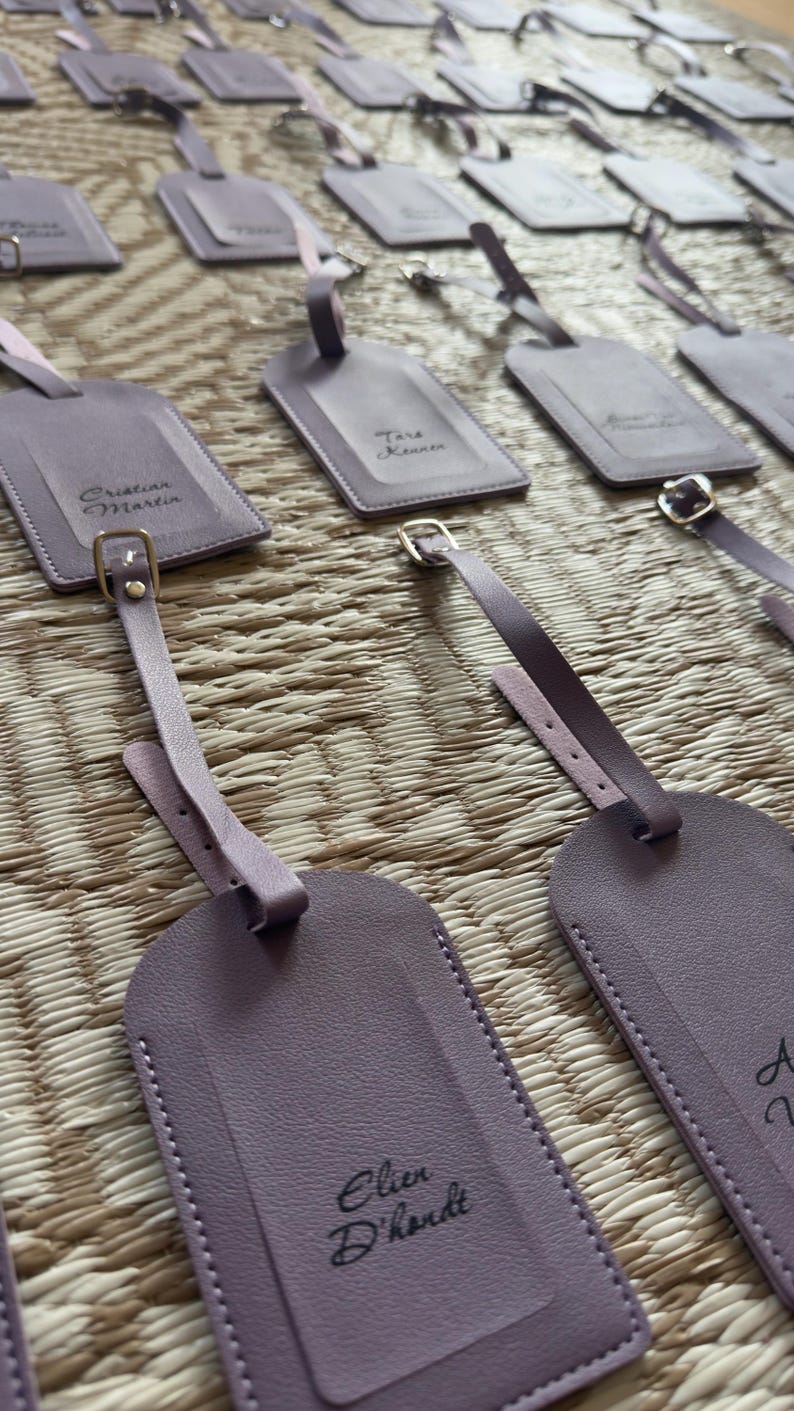 May include: Lavender luggage tags with personalized names in black script. Each tag has a matching strap with a gold-coloured buckle. The tags are arranged on a woven surface.