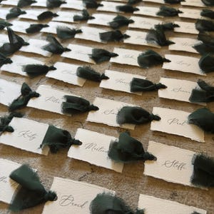 May include: Arrangement of white place cards with dark green ribbon accents. Each card features elegant calligraphy names. The cards are arranged on a textured surface, creating a sophisticated and organized display, perfect for a wedding or event.