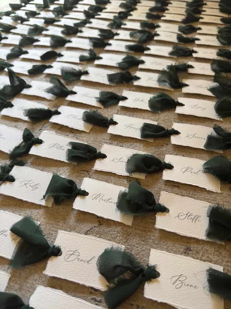May include: Arrangement of white place cards with dark green ribbon accents. Each card features elegant calligraphy names. The cards are arranged on a textured surface, creating a sophisticated and organized display, perfect for a wedding or event.