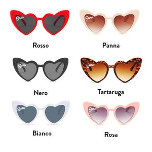 May include: Heart-shaped sunglasses in six colors: red, cream, black, tortoise shell, white, and pink. Each pair has the name "Sara" printed on the upper left lens. The sunglasses have dark lenses and a retro style.