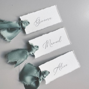 May include: Three white place cards with torn edges, each featuring a name in elegant cursive script: Ginevra, Manuel, and Alice. Each card is tied with a sage green satin ribbon. The background is a solid gray.