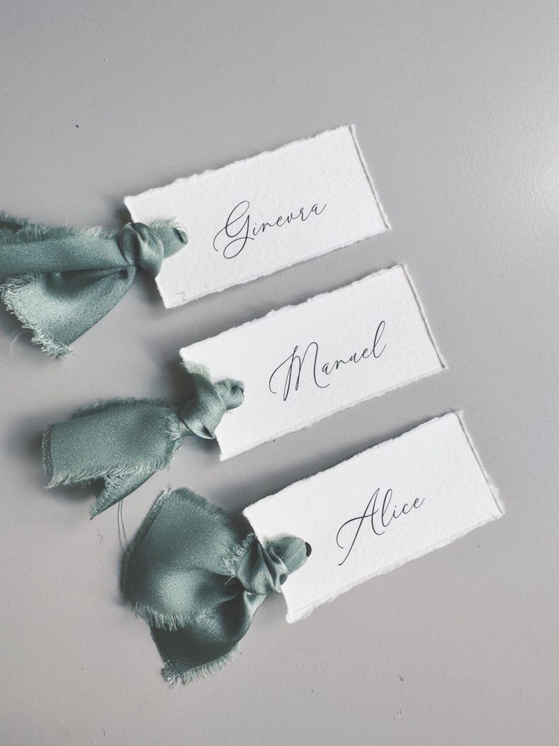 May include: Three white place cards with torn edges, each featuring a name in elegant cursive script: Ginevra, Manuel, and Alice. Each card is tied with a sage green satin ribbon. The background is a solid gray.