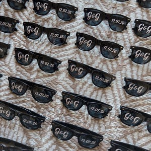 May include: Black sunglasses with the text "G&G" and the date "12.07.26" printed on the lenses. The sunglasses are arranged in a repeating pattern on a textured surface.