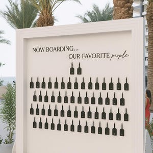 May include: A white wooden sign with the text "NOW BOARDING... OUR FAVORITE people" in black script. The sign features rows of black key-shaped tags, likely for a wedding or event seating arrangement, set against a backdrop of palm trees.