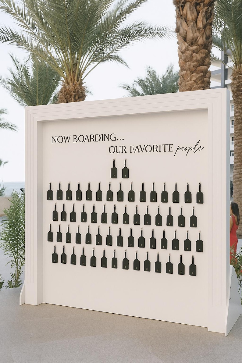 May include: A white wooden sign with the text "NOW BOARDING... OUR FAVORITE people" in black script. The sign features rows of black key-shaped tags, likely for a wedding or event seating arrangement, set against a backdrop of palm trees.