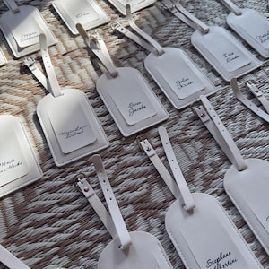 May include: A selection of off-white luggage tags, each with a strap and silver buckle. The tags have a smaller, rectangular section with a name printed in black script. The tags are arranged on a textured, light brown surface.