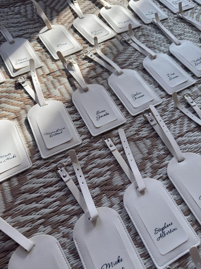 May include: A selection of off-white luggage tags, each with a strap and silver buckle. The tags have a smaller, rectangular section with a name printed in black script. The tags are arranged on a textured, light brown surface.