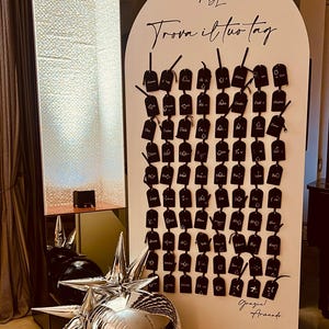 May include: A white sign with the text "Trova il tuo tag" and many black tags with white writing. The sign has the initials "ADL" at the top. Decorative silver star and sphere ornaments are in the foreground.