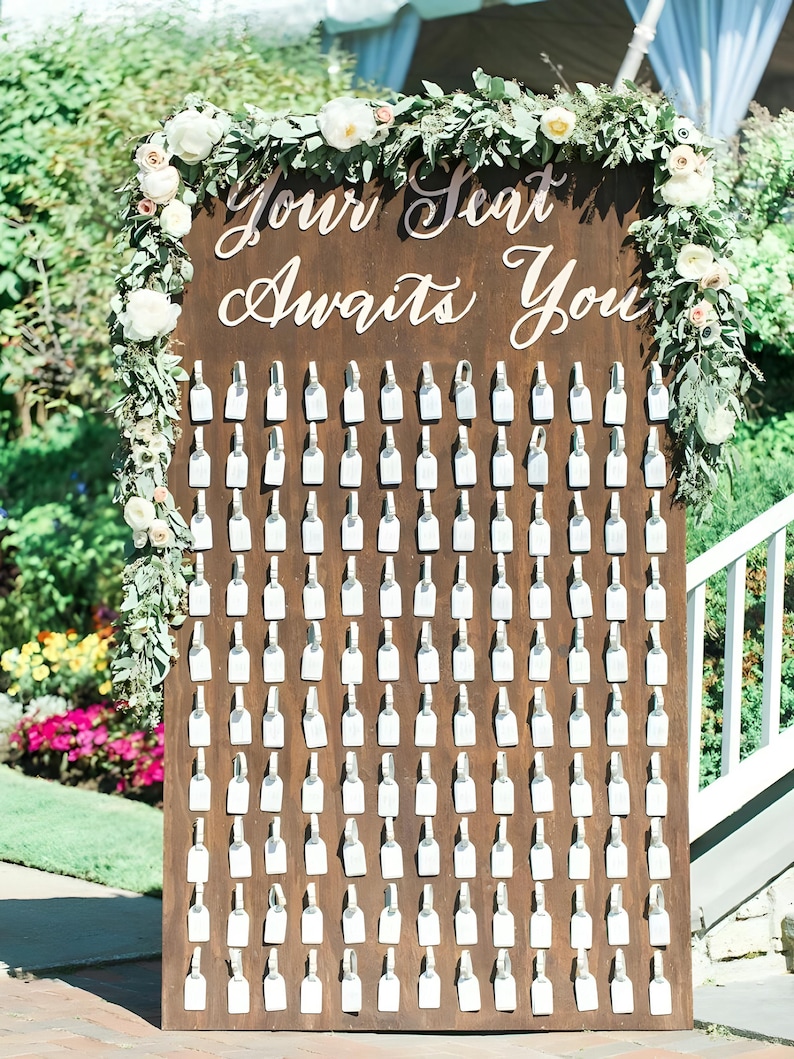 May include: A wooden seating plan with the words "Your Seat Awaits You" in white script. The plan features many small, white, bottle-shaped tags, and is decorated with floral arrangements.