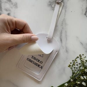 May include: White luggage tag with a silver buckle and a clear window. The tag has the text "TAVOLO ORCHIDEA" printed on a white background. A hand is peeling back a layer of the tag. A small bouquet of white flowers and green leaves is in the corner.