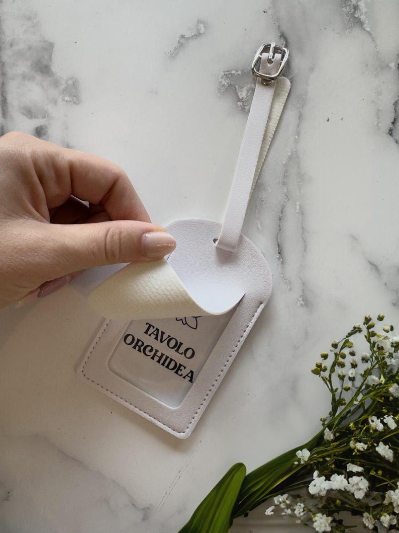 May include: White luggage tag with a silver buckle and a clear window. The tag has the text "TAVOLO ORCHIDEA" printed on a white background. A hand is peeling back a layer of the tag. A small bouquet of white flowers and green leaves is in the corner.