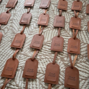 May include: Numerous brown leather luggage tags, each with a rectangular shape and a small top flap. The tags are attached to a strap and display the initials "D&R" and the date "02.05.2026".
