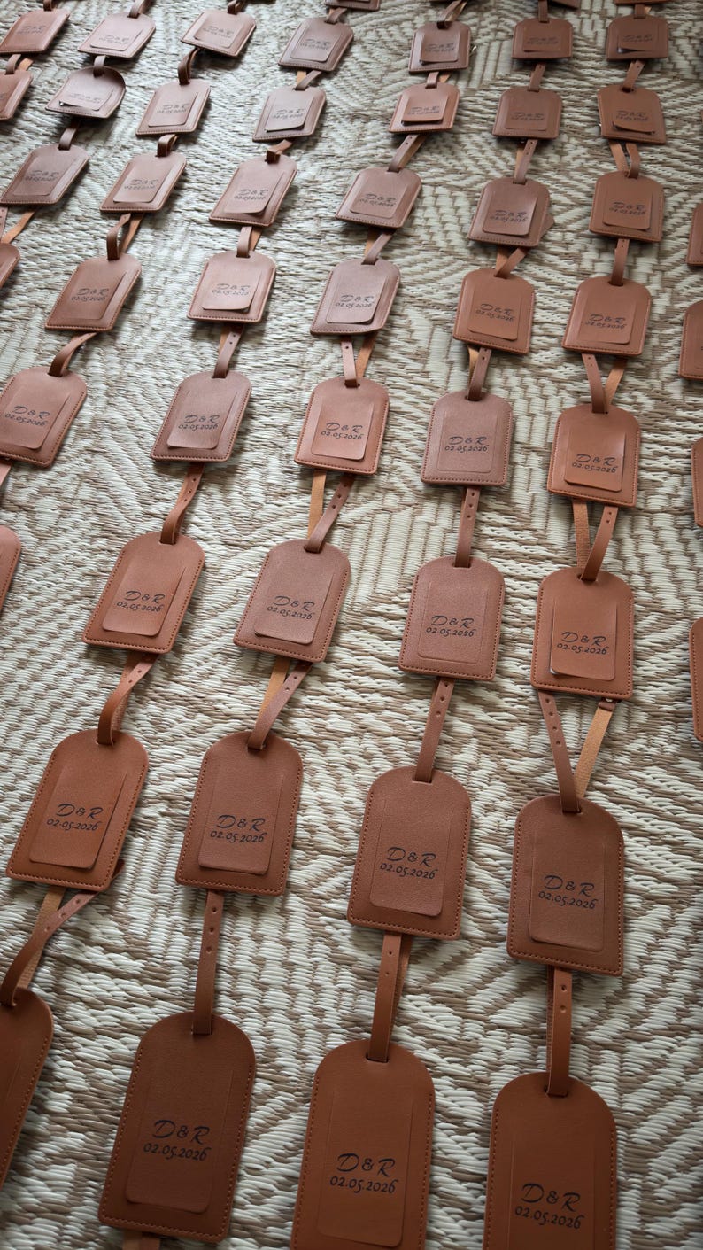 May include: Numerous brown leather luggage tags, each with a rectangular shape and a small top flap. The tags are attached to a strap and display the initials "D&R" and the date "02.05.2026".