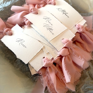 May include: A collection of handmade, off-white paper place cards with torn edges. Each card is tied with a delicate, light pink ribbon. The cards are inscribed with elegant calligraphy in black ink, perfect for weddings or special events.