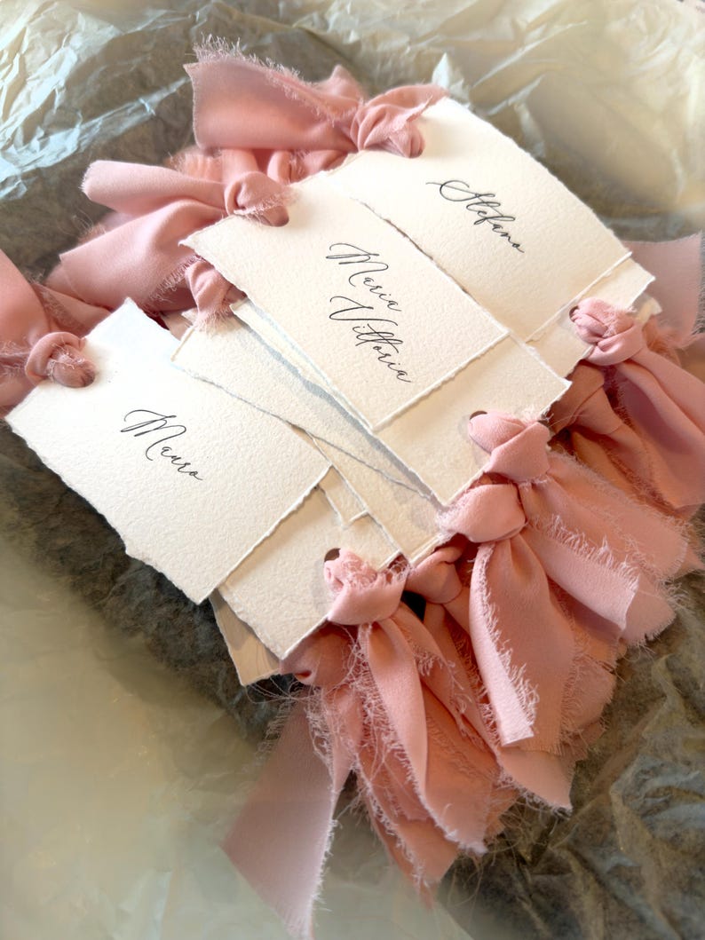 May include: A collection of handmade, off-white paper place cards with torn edges. Each card is tied with a delicate, light pink ribbon. The cards are inscribed with elegant calligraphy in black ink, perfect for weddings or special events.