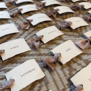 May include: A collection of place cards with names printed in black on textured white paper. Each card is adorned with a light brown ribbon bow. The cards are arranged on a woven surface, creating an elegant display.