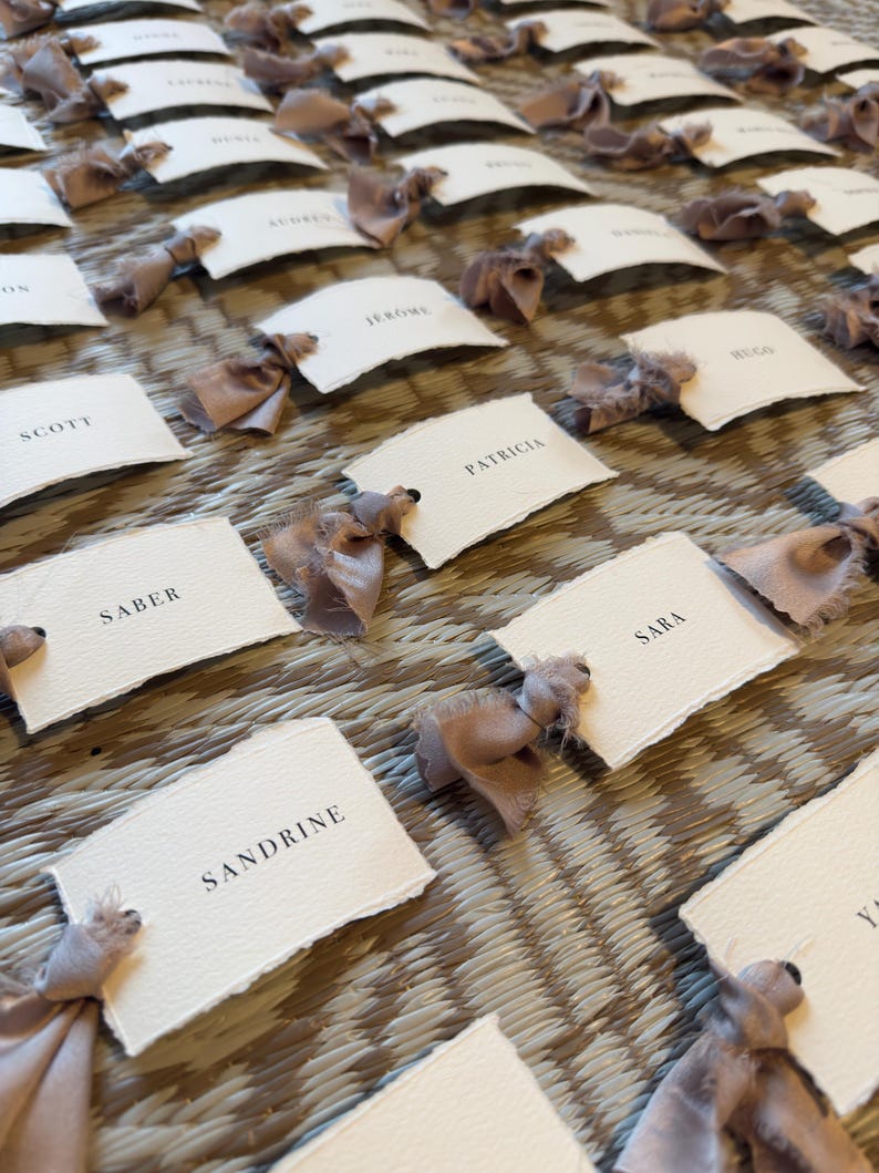 May include: A collection of place cards with names printed in black on textured white paper. Each card is adorned with a light brown ribbon bow. The cards are arranged on a woven surface, creating an elegant display.