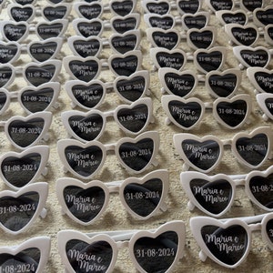 May include: Rows of white heart-shaped sunglasses with black lenses are arranged on a surface. Each pair has the text "Maria e Marco" and the date "31-08-2024" printed on the lenses, likely for a wedding or event.
