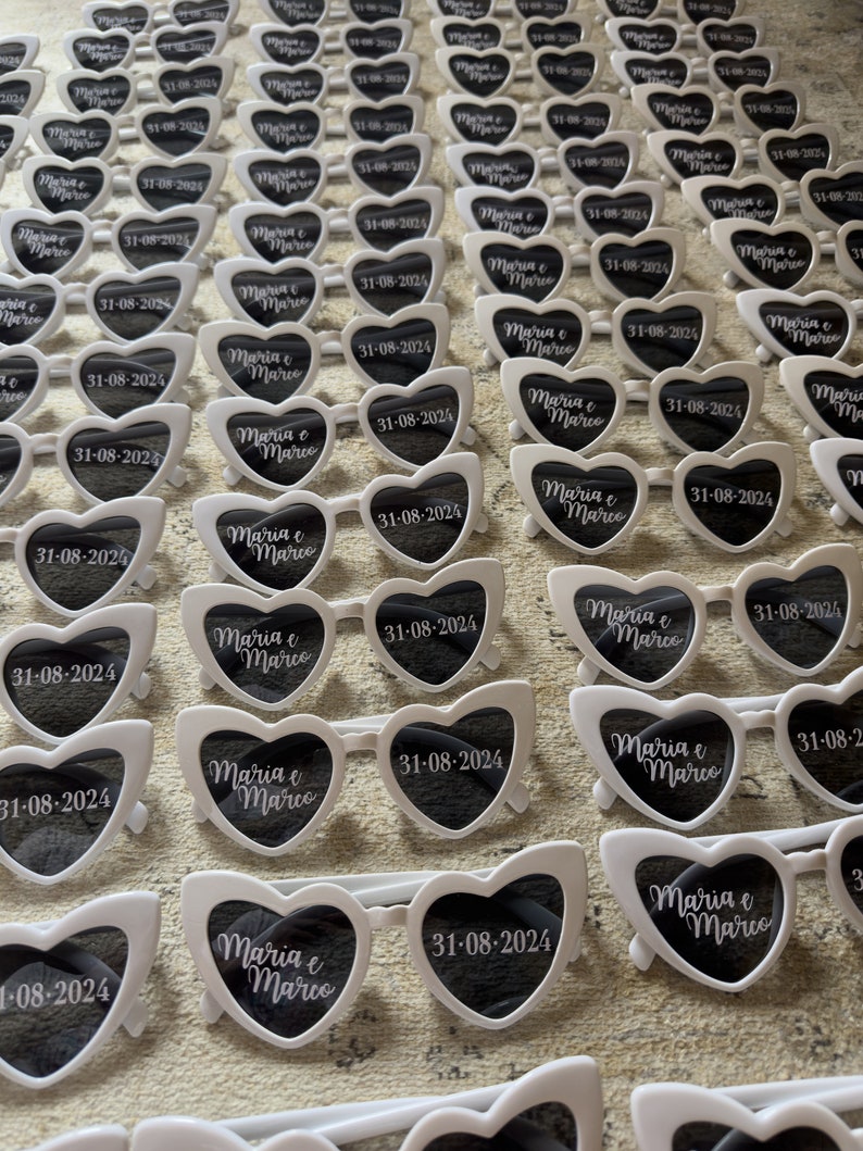 May include: Rows of white heart-shaped sunglasses with black lenses are arranged on a surface. Each pair has the text "Maria e Marco" and the date "31-08-2024" printed on the lenses, likely for a wedding or event.