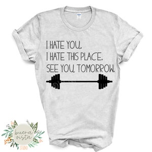 May include: A light gray t-shirt with the text "I HATE YOU. I HATE THIS PLACE. SEE YOU TOMORROW." and a barbell graphic in black. The shirt has rolled-up sleeves and a crew neck.